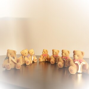 May include: A collection of small, tan teddy bear figurines, each holding a gold letter. Some bears have pink or purple bows. The letters spell out a name. The bears are arranged on a dark surface.