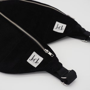 Könnte beinhalten: Schwarze Cord-Bauchtasche mit Reißverschluss und verstellbarem Gurt. Die Tasche hat zwei weiße Etiketten mit dem Text "Jecle Studio" auf jedem.