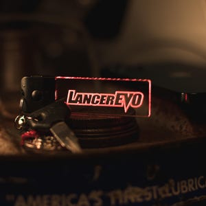 May include: A red acrylic keychain with the text "LancerEvo" in white lettering. The keychain is attached to a black metal ring and a silver key.