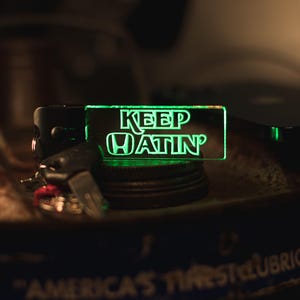 May include: A black key fob with a green neon sign that reads "KEEP HATIN'" in white letters. The sign has a Honda logo. The key fob is resting on a dark surface with keys attached.