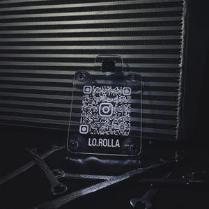 May include: A clear acrylic keychain with a QR code and the Instagram logo. The text "LO.ROLLA" is printed below the QR code. The keychain is displayed in front of a metallic background and tools.