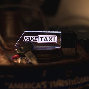 May include: A white acrylic keychain with the words "FAKE TAXI" in black lettering. The keychain is sitting on a dark surface with a key and a small metal object.