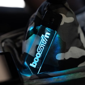 May include: A rectangular, transparent device with the word "boostin" illuminated in blue. The device is attached to a black object, possibly a phone. The background is a camouflage pattern.