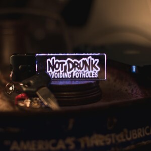 May include: A white acrylic keychain with the text "Not Drunk Avoiding Potholes" in a graffiti-style font. The keychain is illuminated from behind, creating a glowing effect.