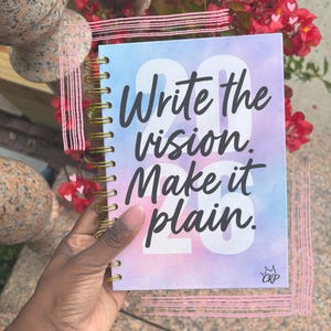 Può includere: Un taccuino a spirale con un design acquerello pastello. La copertina presenta il testo "Write the vision. Make it plain." in nero, con il numero "23" in bianco. Rilegatura a spirale dorata.