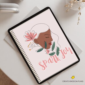 May include: A pink and white spiral bound notebook with a line drawing of a woman's face with flowers and leaves. The text "Spark Joy" is written in pink cursive.