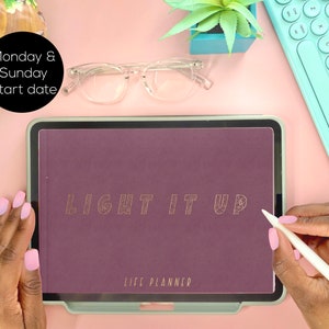 May include: A purple life planner with the words "LIGHT IT UP" in gold lettering on the cover. The planner is on a tablet with a white stylus next to it.