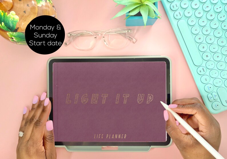 May include: A purple life planner with the words "LIGHT IT UP" in gold lettering on the cover. The planner is on a tablet with a white stylus next to it.