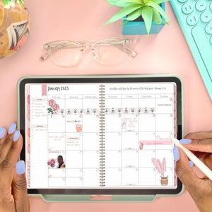 May include: A close-up of a digital planner open to January 2023. The planner has a floral design and is decorated with stickers. The planner is on a pink surface with a keyboard and glasses.