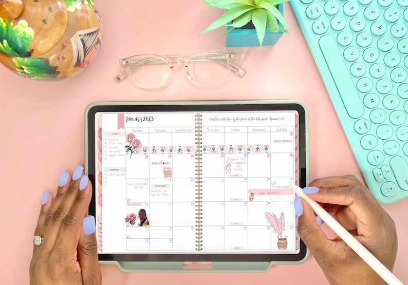 May include: A close-up of a digital planner open to January 2023. The planner has a floral design and is decorated with stickers. The planner is on a pink surface with a keyboard and glasses.