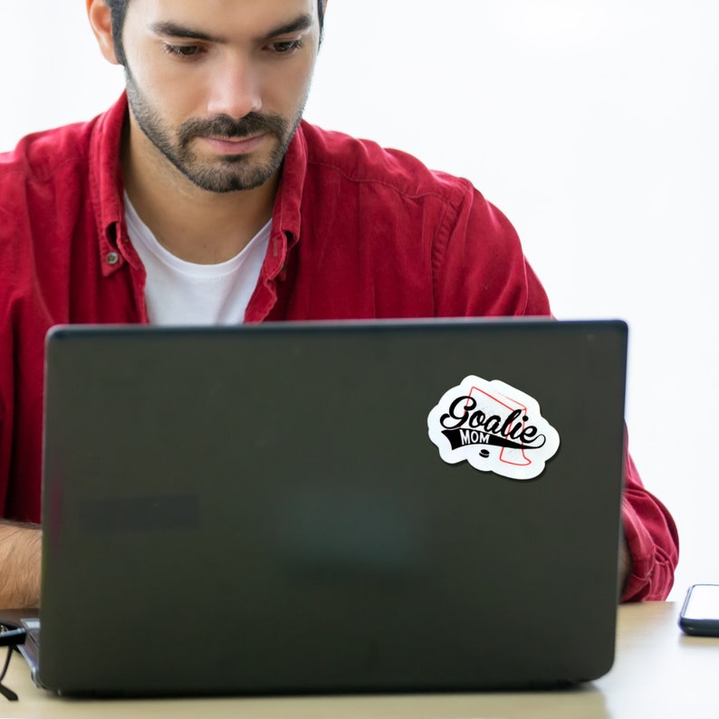 May include: A black laptop computer with a white sticker that says "Goalie Mom" in black text with a red hockey stick graphic.