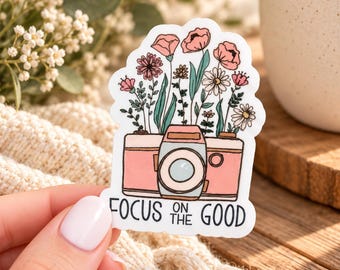 May include: A die-cut sticker featuring a pink camera with a bouquet of various pink and white flowers emerging from the top. The text "FOCUS ON THE GOOD" is printed in black capital letters at the bottom of the sticker design.
