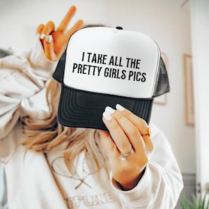 May include: A black and white trucker hat with the text "I TAKE ALL THE PRETTY GIRLS PICS" in bold, black letters. The hat has a white front panel and a black mesh back. The brim is black.