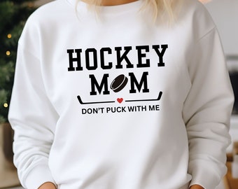 May include: White sweatshirt with black text that reads "HOCKEY MOM" with a hockey puck graphic between the words "MOM". Below the text is a hockey stick graphic and the text "DON'T PUCK WITH ME".