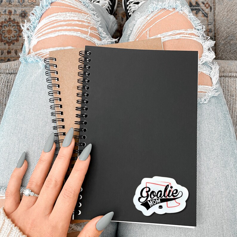 May include: A black spiral bound notebook with a white sticker that says "Goalie Mom" on the front. The notebook is sitting on a person's lap.