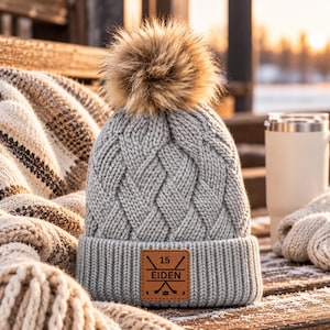 May include: A light grey cable-knit beanie featuring a large, fluffy brown faux fur pom-pom on top. The cuff displays a square tan leather patch engraved with the number 15, the name EIDEN, and a graphic of two crossed hockey sticks.