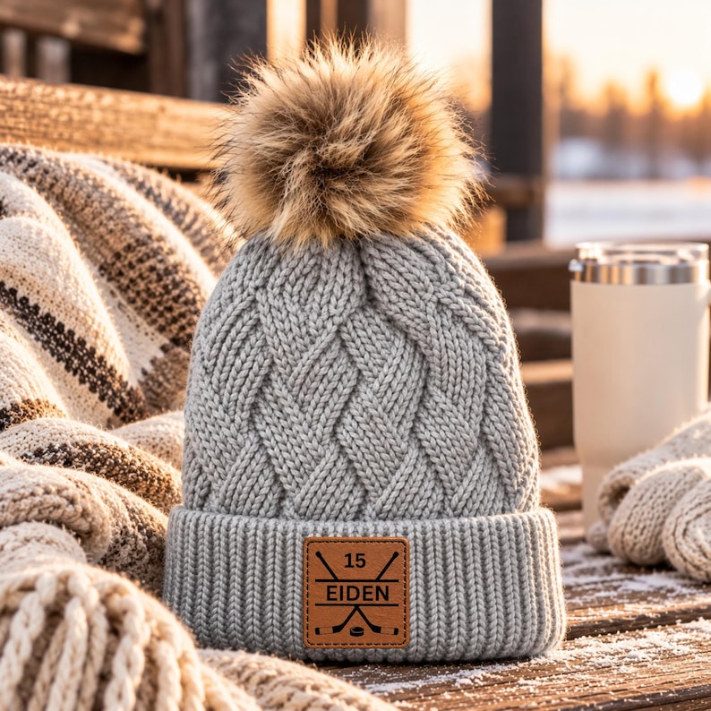 May include: A light grey cable-knit beanie featuring a large, fluffy brown faux fur pom-pom on top. The cuff displays a square tan leather patch engraved with the number 15, the name EIDEN, and a graphic of two crossed hockey sticks.