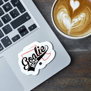 May include: A white sticker with black and red text that says "Goalie Mom" with a hockey net and puck graphic. The sticker is on a silver laptop.