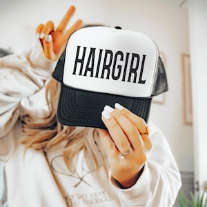 May include: A black and white trucker hat with the word "HAIRGIRL" printed in bold, black letters. The hat has a white front panel and a black brim and mesh back. The hat is being held up by a person.