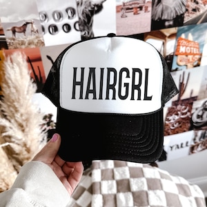 May include: Black and white trucker hat with the word "HAIRGRL" printed in bold, black letters on the front. The hat has a white front panel and a black brim and mesh back. The hat is a classic style.