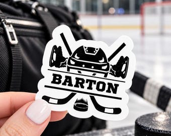 May include: A white die-cut sticker featuring a black graphic of a hockey helmet centered between two crossed hockey sticks. Below the helmet, the name BARTON is printed in bold black capital letters, with a hockey puck positioned at the bottom.