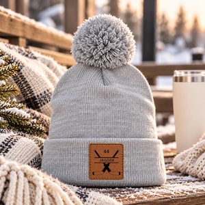 May include: A light grey knit beanie with a matching pom-pom on top. The front features a square brown leather patch with a graphic of two crossed hockey sticks, the number 44, and the name JAGGER printed in black ink.