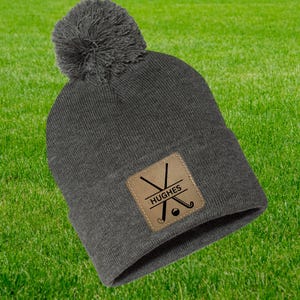May include: A charcoal grey beanie hat with a matching pom-pom. It has a brown square patch with a golf design and the name "HUGHES". The background is green grass.