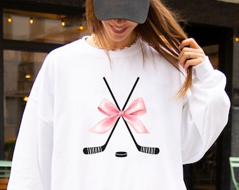 May include: White sweatshirt with a pink bow tied around two crossed hockey sticks and a black hockey puck.