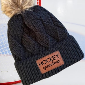 May include: Black knitted beanie with a faux fur pom pom and a brown leather patch that says "HOCKEY grandma".