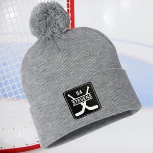 May include: A gray knit beanie with a pom-pom on top. The beanie features a black square patch with the number "54", the word "STEVENS", and crossed hockey sticks.