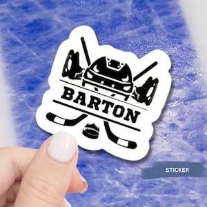 Puede incluir: Una pegatina blanca con un casco de hockey negro, palos de hockey cruzados y el nombre "BARTON" impreso en una pancarta negra.