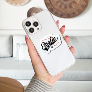 May include: White iPhone case with a black and red sticker that says "Goalie Mom" on it.