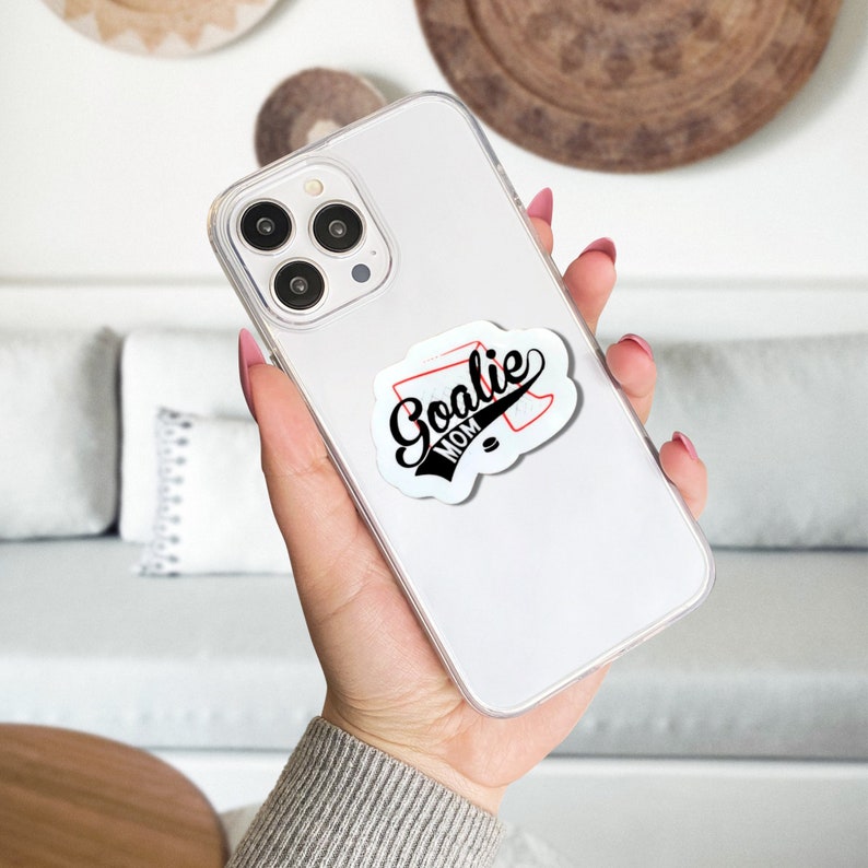 May include: White iPhone case with a black and red sticker that says "Goalie Mom" on it.