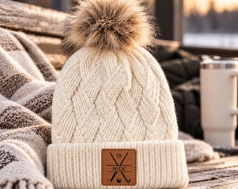 May include: A cream-coloured knit beanie with a cable-knit pattern and a large faux fur pom-pom on top. The beanie features a folded cuff with a square brown leather patch engraved with the number 15, the name NICK, and hockey sticks.
