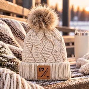 May include: A cream-colored cable knit beanie featuring a large faux fur pom-pom on top. The cuff displays a square brown leather patch engraved with the number 17, a hockey stick graphic, and the text WALKER, along with a small hockey puck.