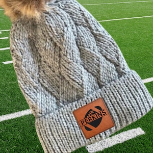 May include: Grey knitted beanie with a faux fur pom pom and a brown leather patch featuring a football graphic and the name "PERKINS" in white text.