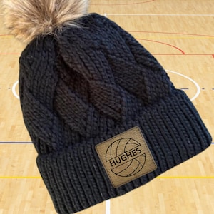May include: Black knitted beanie with a faux fur pom pom and a brown leather patch with a volleyball graphic and the text "HUGHES".