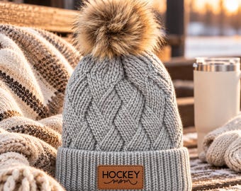 May include: A light grey knit beanie featuring a thick cable knit pattern and a large, fluffy brown faux fur pom-pom on top. The beanie has a wide ribbed cuff with a rectangular brown leather patch centred on the front, embossed with the text HOCKEY mom.