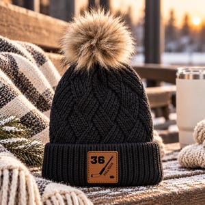 May include: A black knit beanie with a cable-knit pattern and a large faux fur pom-pom on top. The cuff features a square tan leather patch embossed with the number 36, a hockey stick, a hockey puck, and the text HILLIGUS.