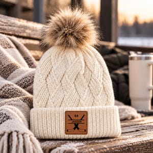 May include: A cream-colored cable-knit beanie with a large faux fur pom-pom on top. The cuff features a square brown leather patch with the text 44 and JAGGER, along with a graphic of two crossed hockey sticks in black ink.
