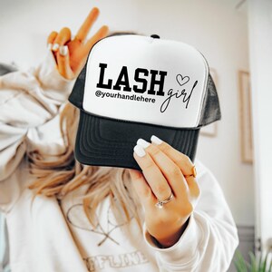 May include: A black and white trucker hat with the text "LASH girl" and the Instagram handle "@yourhandlehere" printed on the front. The hat has a white front panel and a black brim and mesh back.
