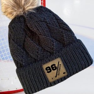 May include: Black cable knit beanie with a faux fur pom pom and a brown leather patch with the text "96" and "SCHNECK" in black.
