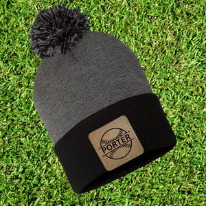 Può includere: Un berretto grigio e nero con un pompon scuro. Il berretto presenta una toppa in pelle con un disegno di baseball e il nome "PORTER". Il berretto è su uno sfondo di erba verde.