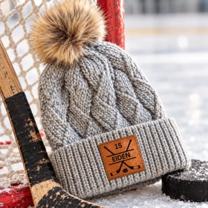 May include: A grey cable-knit beanie with a large brown faux fur pom-pom on top. The beanie features a tan leather patch on the ribbed cuff, engraved with the number 15, the name EIDEN, and a graphic of two crossed hockey sticks and a puck.