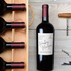 Pode incluir: Uma garrafa de vinho tinto com um rótulo que diz "Pairs well with a great Hockey Manager. Thank you for all you do." O rótulo também tem um par de tacos de hóquei cruzados.