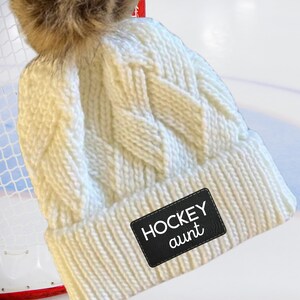 May include: White knitted beanie with a faux fur pom pom and a black leather patch that says "HOCKEY aunt".