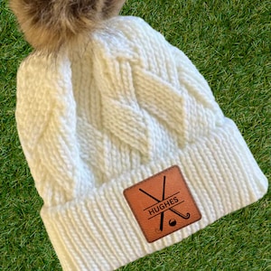 May include: White knitted beanie with a faux fur pom pom and a brown leather patch with the name "HUGHES" and a hockey stick graphic.