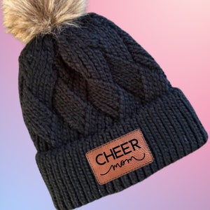 May include: Black knitted beanie with a faux fur pom pom and a brown leather patch that says "CHEER mom".