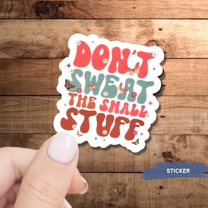 May include: A white sticker with the text "Don't sweat the small stuff" in a retro font with colorful accents and butterflies. The sticker is held by a hand.