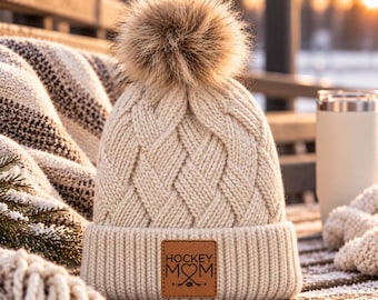 May include: A cream-coloured cable knit beanie featuring a large faux fur pom-pom on top and a square brown leather patch on the cuff. The patch is engraved with the text HOCKEY MOM and a graphic of two crossed hockey sticks with a heart.